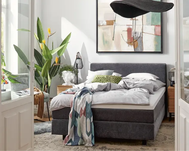 Modern eingerichtetes Schlafzimmer mit hochwertigem Polsterbett in Grau, gemütlicher Bettwäsche, Holznachttischen und Pflanzen. Stilvolle Schlafzimmereinrichtung mit Fokus auf Komfort, Design und wohnlicher Atmosphäre.